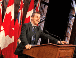 A man standing at a podium with multiple canadian flags behind him