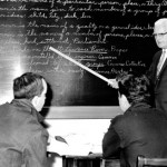 A man pointing at a board with writing while looking at two people